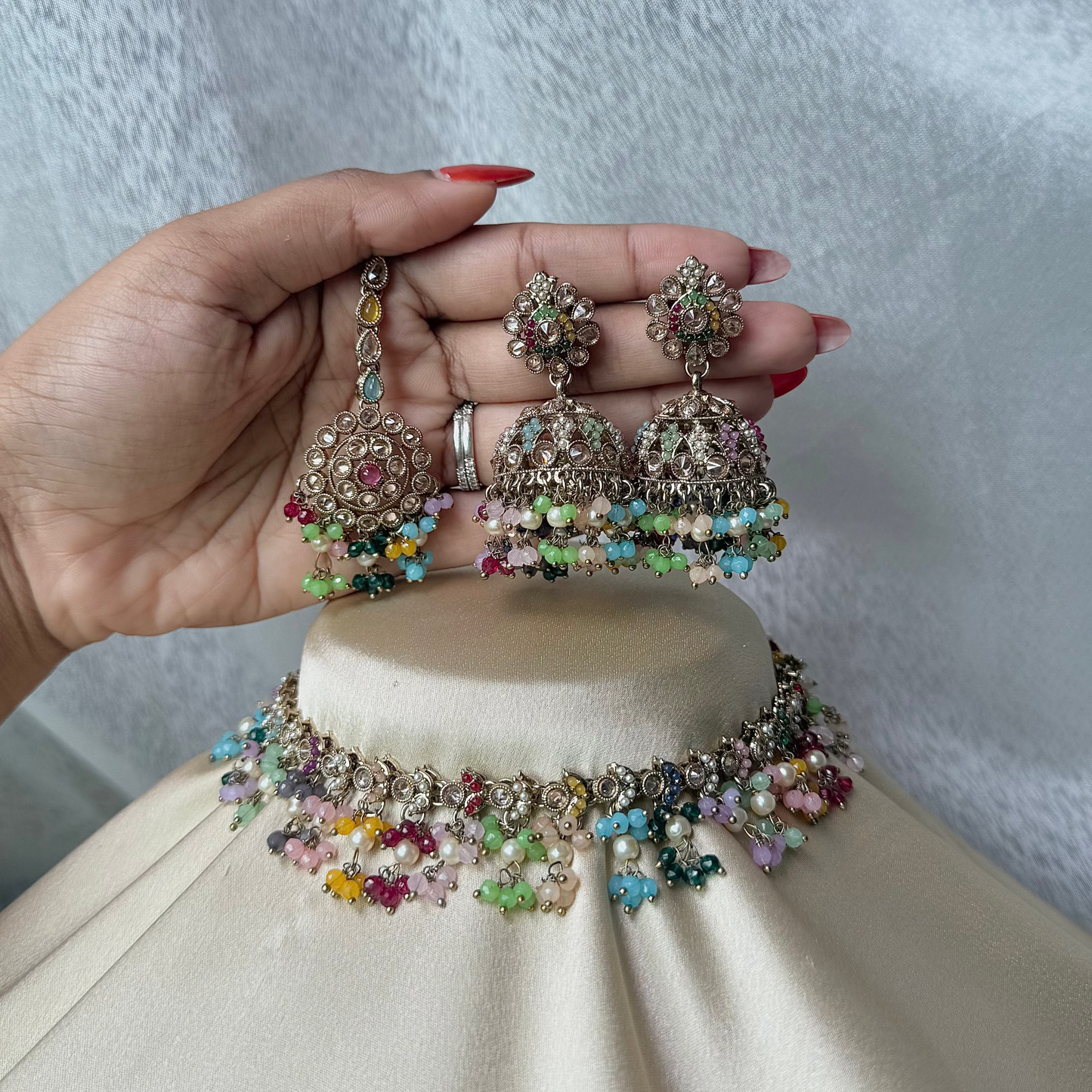 Colorful jewelry set on a small round stand held by a hand with red nail polish against a light background.